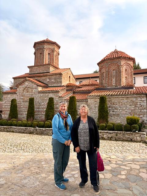       Two women posing happily in front of a stone-built Byzantine monastery with red tiled roofs.
  