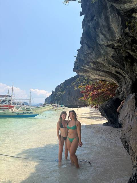       Two women in swimsuits stand on a white sand beach near outrigger boats and limestone cliffs.
  