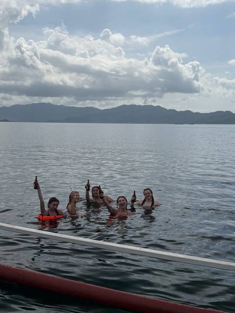       Five friends waist-deep in the sea raising beer bottles with hazy mountains in the background.
  
