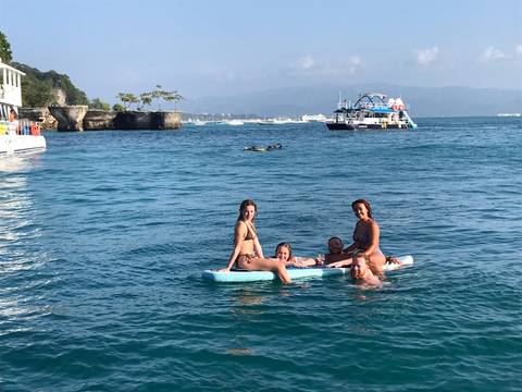       Small group lounging on a paddleboard in turquoise sea near rocky coastline and boats.
  