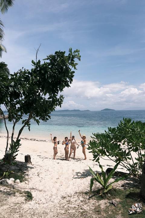       Four women celebrating with drinks on a white-sand tropical beach framed by leafy trees and distant islands.
  