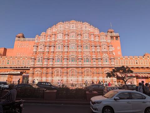      Front façade of Jaipur’s pink Hawa Mahal glowing under clear blue skies with street life below.
  