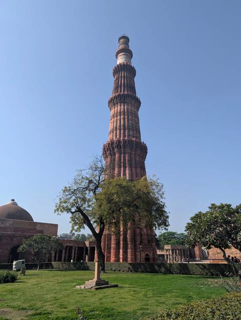       Towering Qutub Minar rising above surrounding trees against a clear blue Delhi sky.
  