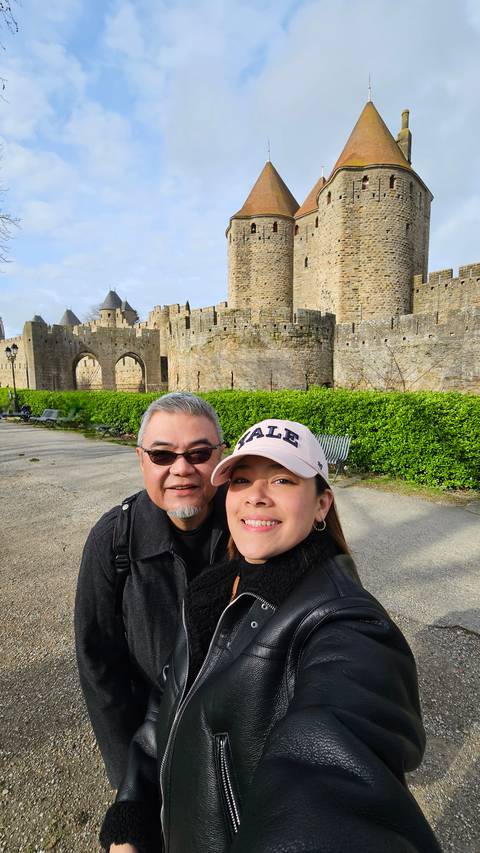       Smiling couple taking a selfie with the medieval walls and towers of Carcassonne behind.
  