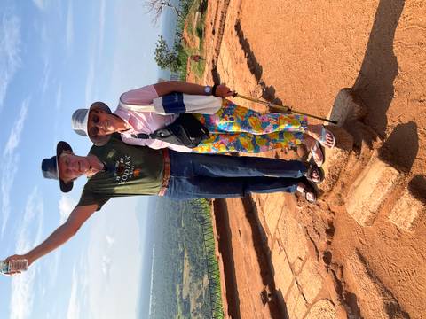       Happy couple posing triumphantly on an ochre rock platform high above lush plains.
  
