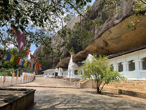       White façade of Dambulla cave temple nestling under a vast cliff overhang adorned with flags.
  