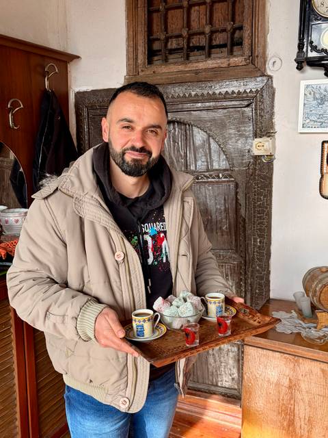       Local host holding a plate of Turkish delight and tiny coffee cup inside a rustic house.
  
