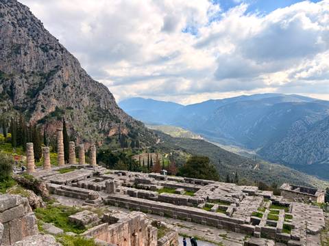       Panoramic view of Delphi ruins with pine-covered mountains and dramatic clouds beyond.
  