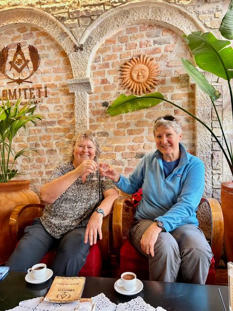       Two women clinking tiny shot glasses in a cozy bar with exposed brick walls.
  