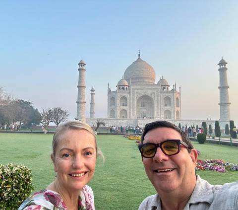       Couple posing for a selfie with the iconic Taj Mahal and its minarets rising behind them in soft morning light.
  