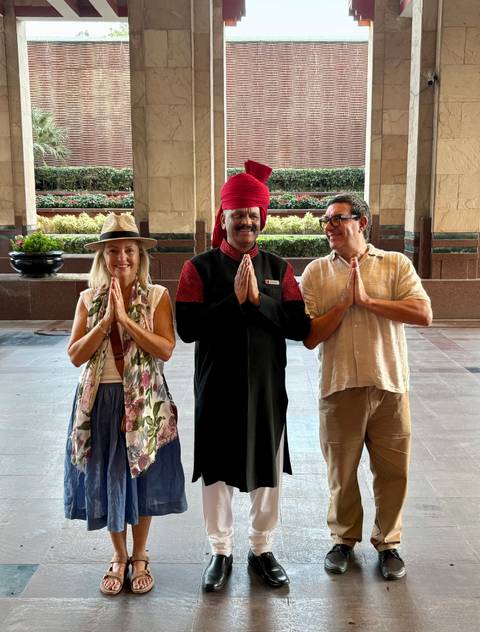       Couple standing with a traditional-dressed doorman inside a grand hotel lobby, all greeting with a namaste gesture.
  