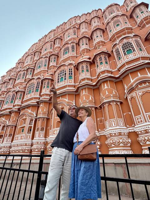       Couple pointing up at the ornate pink façade of Jaipur’s Hawa Mahal while taking a selfie style shot.
  