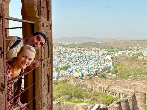       Playful couple leaning from a fortress window overlooking the blue houses of Jodhpur spread across the valley below.
  