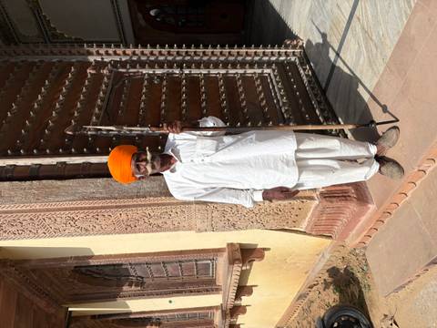       Portrait of a Rajasthani guard in white traditional attire and bright orange turban standing by a carved wooden doorway.
  