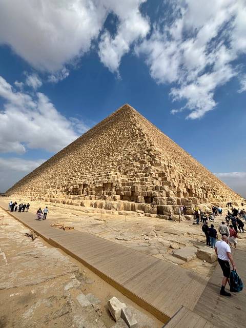       Dramatic low-angle view of the Great Pyramid of Giza with tiny visitors at its base against a blue sky with clouds
  