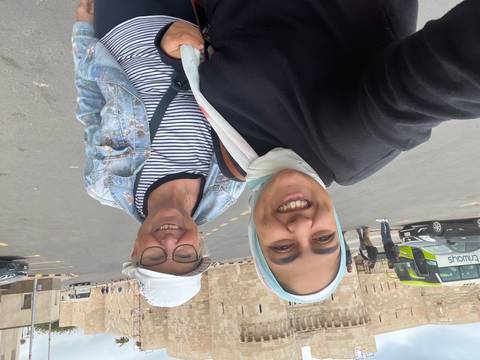       Two smiling travellers take a selfie in front of historic stone walls and parked tour buses.
  