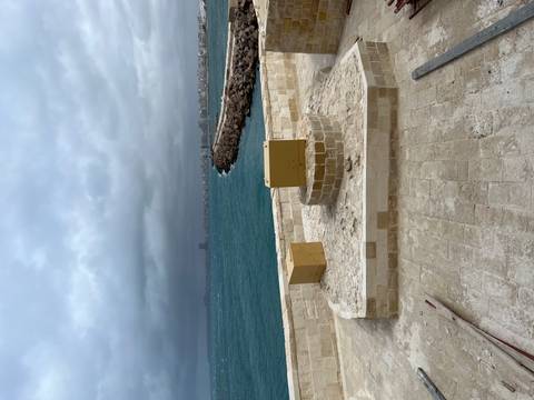       Overcast seafront terrace overlooking a breakwater and choppy turquoise Mediterranean waters under grey skies.
  