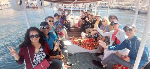       Cheerful tour group seated on an open boat cruising a calm river with city docks in the background, all flashing peace signs.
  