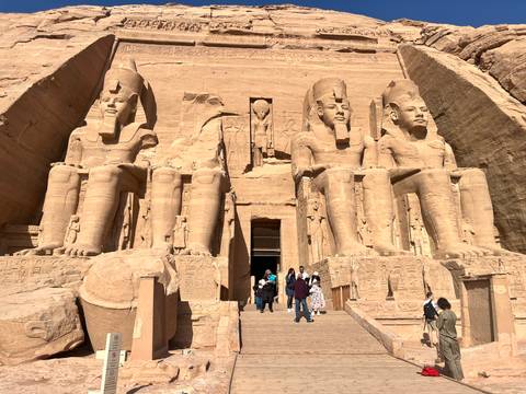       Grand façade of Abu Simbel with four colossal seated statues of Ramses II towering above visitors on the stone steps.
  
