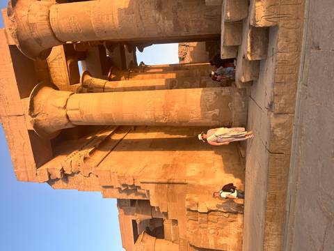       Golden sunlight illuminates towering sandstone columns of an Egyptian temple as visitors stroll between them.
  