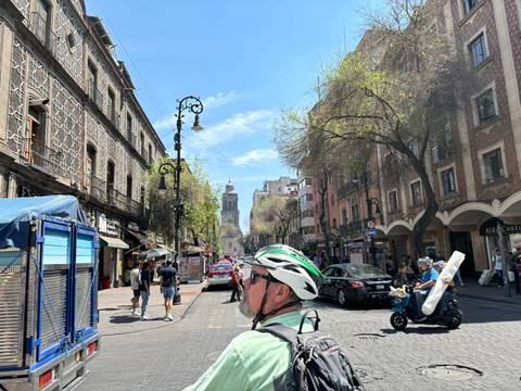       Busy historic street with colonial buildings, pedestrians and a cyclist wearing a helmet in bright sunshine.
  