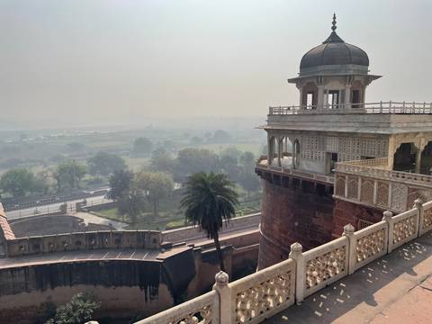      View from Agra Fort ramparts overlooking green plains and historic sandstone towers in morning haze.
  