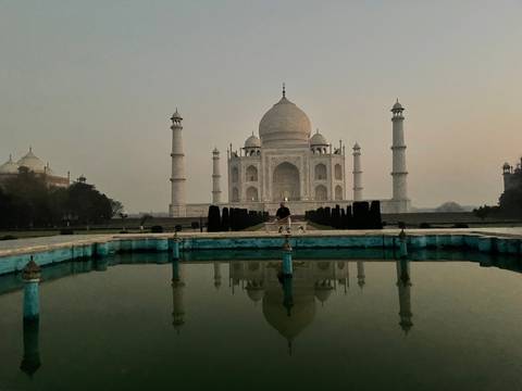       Iconic Taj Mahal reflected in a still pool at dawn with a lone visitor seated on the central platform.
  