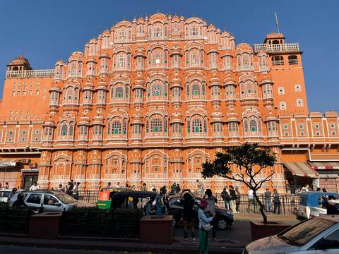       The ornate orange facade of Jaipur's Hawa Mahal with bustling street traffic and pedestrians below.
  