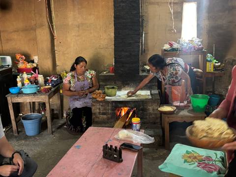       Two indigenous women prepare tortillas over a wood-fired hearth inside a rustic kitchen.
  