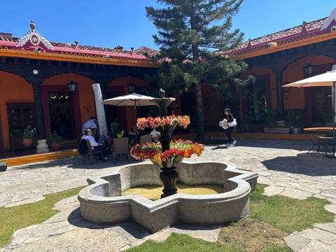       Sunny colonial courtyard with a tiered stone fountain, diners under umbrellas, and vivid orange walls.
  