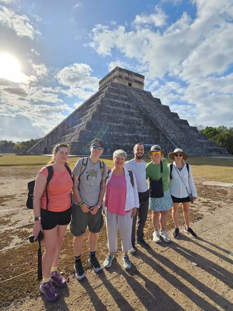      Six travellers smile in front of the massive stepped pyramid at Chichén Itzá on a bright day.
  