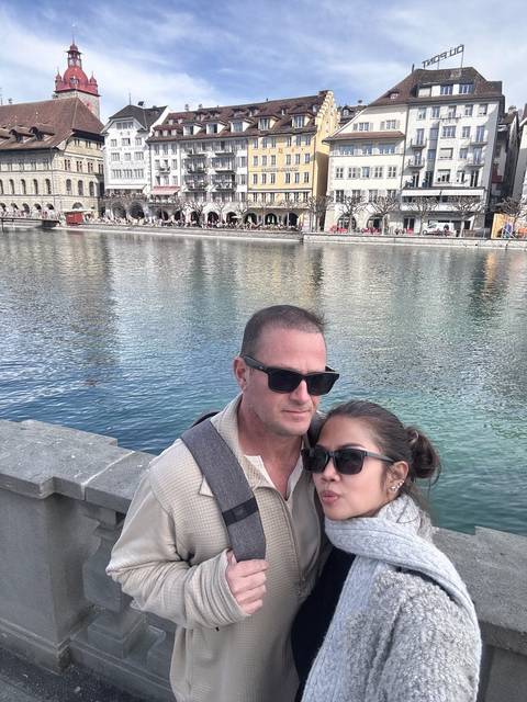       Selfie of a couple wearing sunglasses beside a tranquil riverside quay with arcaded buildings.
  