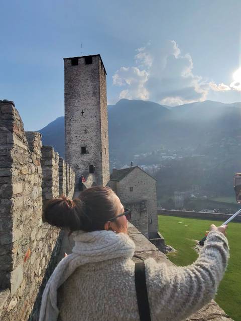       Woman looking out over a medieval stone castle wall with tower and mountains in background
  