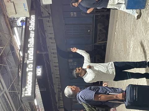      Guide gestures toward illuminated sign reading 'Welcome to Tundla Railway Station' as traveler looks on.
  