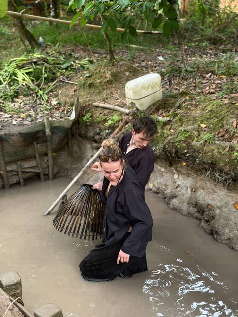       Two travelers wearing dark clothing search for fish with a bamboo basket trap in a muddy canal.
  