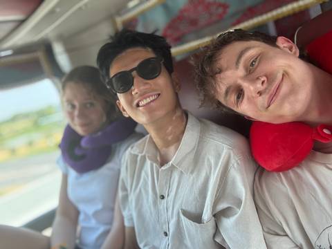       Three young friends take a relaxed selfie on a bus, neck pillows ready for travel.
  