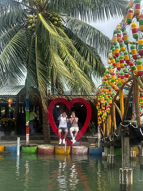       Two friends pose inside a red heart-shaped frame beneath palm trees at a riverside café.
  