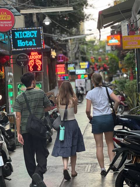      Travelers stroll a neon-lit alley lined with motorbikes, bars and bright signage at night.
  