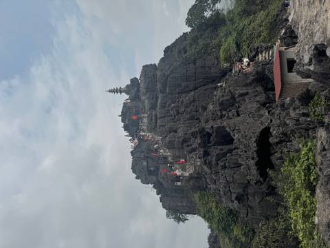       Steep stone steps lead up a dark karst peak topped by a pagoda, dotted with climbers carrying flags.
  