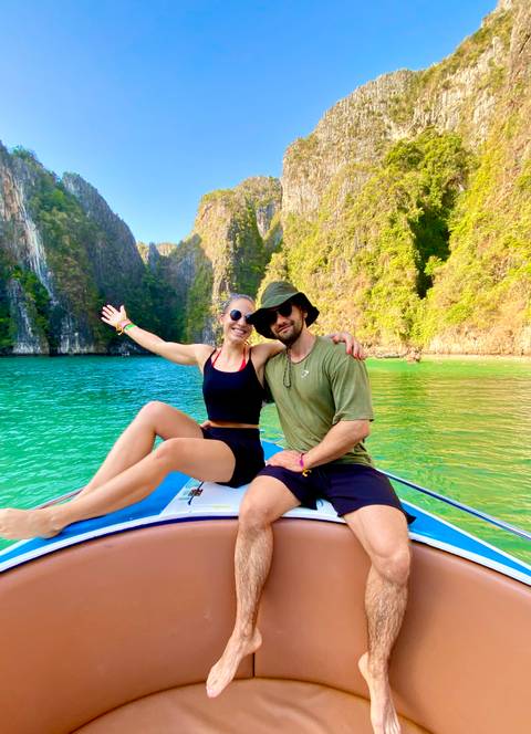       Smiling couple sitting on the bow of a speedboat in turquoise water surrounded by limestone cliffs.
  