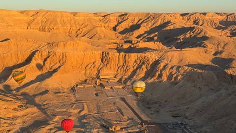       Hot-air balloons drift over the dramatic orange canyons and tomb entrances of the Valley of the Kings at sunrise
  