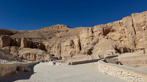       Broad gravel path leading toward rugged cliffs and tomb entrances in the Valley of the Kings against deep blue sky
  