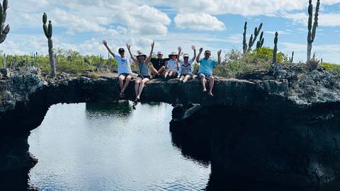       Group of travellers sit with arms raised on a natural lava bridge above clear water surrounded by cactus in the Galápagos.
  