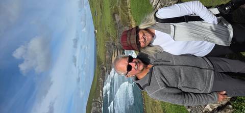       Smiling older couple posing in front of rugged coastal cliffs and crashing waves on a bright day.
  