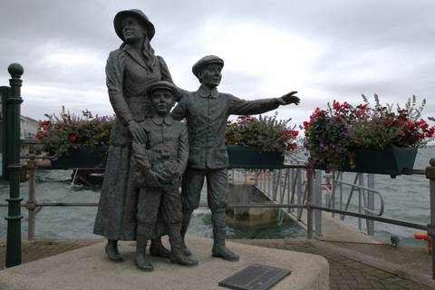       Bronze emigrant family statue on a waterfront promenade decorated with colorful flower boxes on a cloudy day.
  