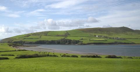       Rolling green patchwork hills meeting a calm bay under blue skies with scattered clouds.
  