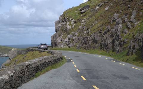       Winding coastal road carved into rocky cliffs with a single parked car and ocean views.
  