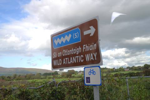       Bilingual road sign marking the Wild Atlantic Way cycling route set against pastoral countryside.
  