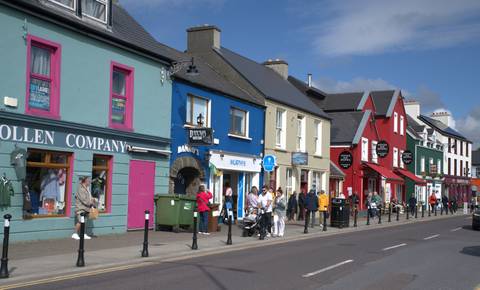       Colorful row of shops on a lively Irish town street with tourists browsing and chatting under blue skies.
  