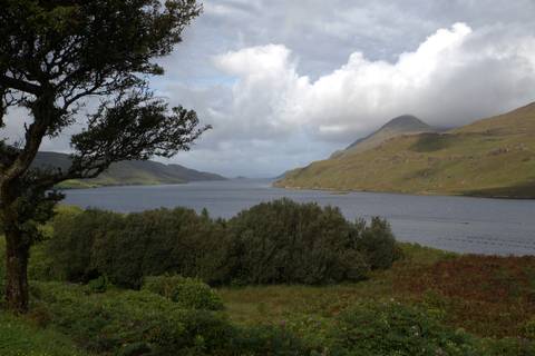       Wide lake inlet surrounded by green hills and a partly shrouded peak under broken clouds.
  